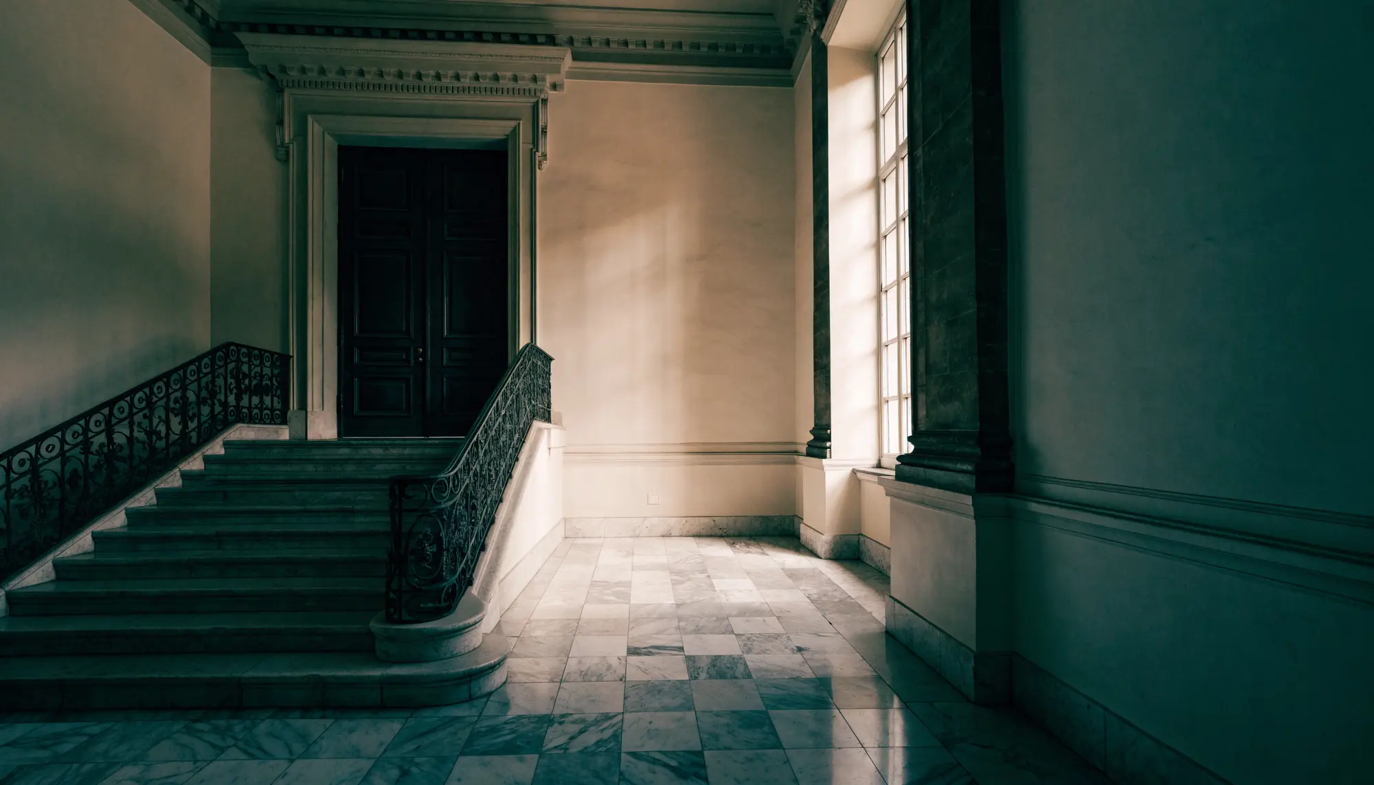 Hall d'entrée en lumière naturelle : escalier de pierre, rampe en fer forgé et hautes portes à panneaux.